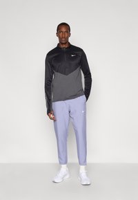 Czarny i szary sportowy top Nike z długim rękawem i ćwierć zamkiem, w połączeniu z jasnofioletowymi dopasowanymi spodniami i białymi sneakersami.