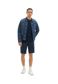 Jeansjacke mit klassischem Schnitt, dunkelblauer Farbe, kombiniert mit einem marineblauen T-Shirt und dunklen Shorts. Weiße Sneaker runden das Outfit ab.