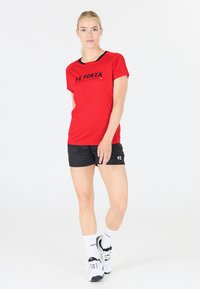 Röd sporttröja med svarta accenter och logotyp, tillsammans med svarta shorts. Modellen har på sig vita strumpor och svarta sportskor, stående på en vit bakgrund.