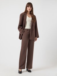 Καφέ κοστούμι προσαρμοσμένο, με σακάκι διπλόστενο και παντελόνι με ίσια γραμμή· συνδυάζεται με κρεμ τοπ και μαύρα παπούτσια με τακούνι.