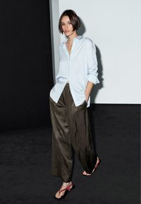 Lichtblauwe overhemd met knopen, oversized pasvorm, opgerolde mouwen, gecombineerd met donkergroene wijde broek en rode platte sandalen. Minimalistische achtergrond.