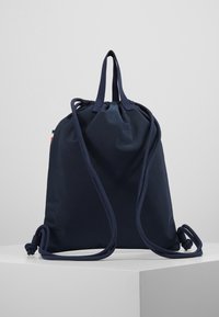 Mochila de cuerdas de color azul marino con dos correas para los hombros y un asa de transporte en la parte superior, erguida sobre una plataforma blanca.