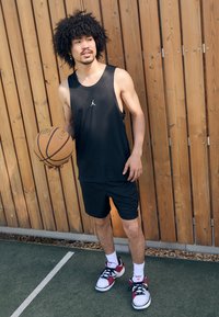 Débardeur noir sans manches avec un logo blanc, short noir assorti et chaussures de sport rouges et blanches. Tenu un ballon de basket, debout sur un terrain.