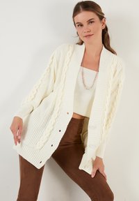 Cárdigan blanco grueso de punto con patrón trenzado, de frente abierto y grandes botones; mangas texturizadas y dobladillo acanalado. Lleva puesto sobre una blusa clara y pantalones marrones.