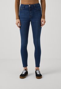 Calças jeans de cintura alta, feitas de tecido denim, com um corte slim. Combinadas com sapatos pretos e uma blusa de manga longa castanha.