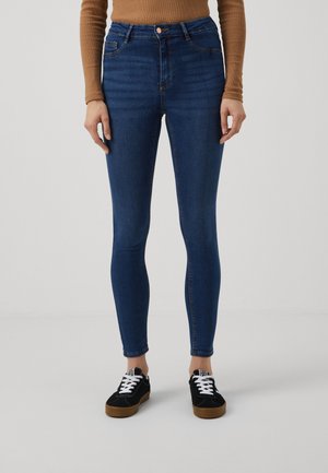Hoge taille blauwe jeans van denimstof, met een slim fit. Gecombineerd met zwarte schoenen en een bruin langmouwen shirt.