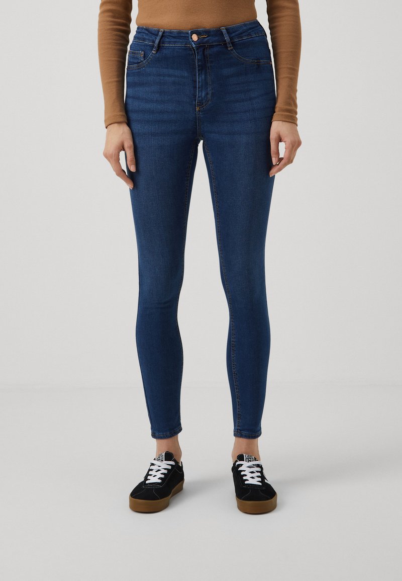 Calças jeans de cintura alta, feitas de tecido denim, com um corte slim. Combinadas com sapatos pretos e uma blusa de manga longa castanha.