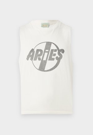 Homme portant des lunettes de soleil noires, un débardeur blanc avec le logo "ARIES", un pantalon noir ample et des chaussures marron, debout devant un fond uni.