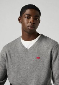 Szary sweter w serek wykonany z miękkiego dzianiny, z małym czerwonym logo na piersi. Nosi się go na białej koszuli.