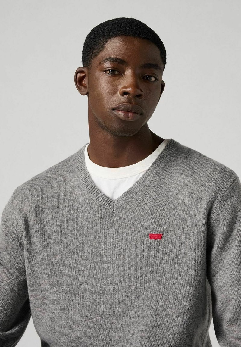 Suéter gris de cuello en V hecho de material de punto suave, con un pequeño logo rojo en el pecho. Usado sobre una camisa blanca.