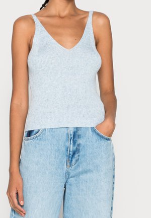 Kobieta ubrana w jasnoniebieski prążkowany, dzianinowy top bez rękawów z dekoltem w serek oraz jasnoniebieskie dżinsy z wysokim stanem, z jedną ręką w kieszeni.