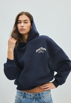 Ung kvinde med langt brunt hår iført en marineblå hættetrøje med teksten "New York Chill Take it easy" og blå jeans mod en ensfarvet baggrund.