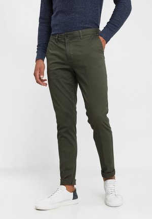 Mężczyzna w ciemnozielonych dopasowanych spodniach, granatowej bluzce z długim rękawem, białych tenisówkach, stojący z jedną ręką w kieszeni.