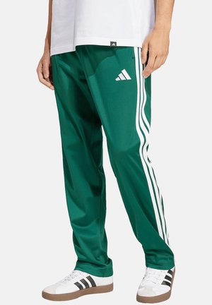 Osoba nosząca zielone spodnie dresowe Adidas z białymi paskami oraz białe trampki z czarnymi paskami na jednolitym jasnym tle.