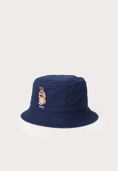 Marineblå bøttehat lavet af stof, prydet med en broderet teddy bjørn og "POLO" logoet foran. Rund, bredskygget form.