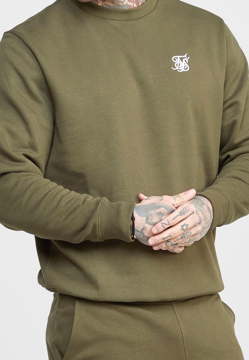 Olivgrön sweatshirt med rundad hals och ribbade ärmslut. Har en liten vit logotyp på vänster bröst. Mjuk, strukturerad tyg.