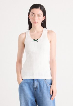 White ribbed tank top with a green bow accent at the neckline. Features a subtle woven pattern and thin straps. Paired with blue denim jeans.