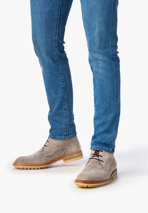 Botas de ante gris claro con puntera redondeada, cordones marrones y suela beige, combinadas con jeans azules. Las botas tienen una superficie texturizada y un agarre de goma.