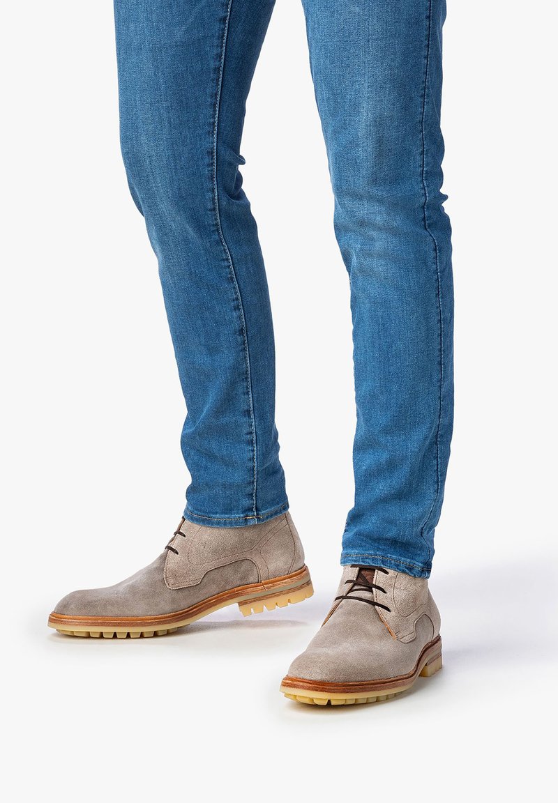 Botas de ante gris claro con puntera redondeada, cordones marrones y suela beige, combinadas con jeans azules. Las botas tienen una superficie texturizada y un agarre de goma.