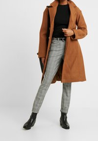Brown, long wool coat over a black top, paired with gray plaid trousers and black ankle boots. Features a belted waist and smooth textures.