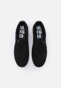 Černé slip-on boty vyrobené z plátna, s hladkým povrchem, minimalistickým designem a bílým logem na stélce. Doplněny malou vlajkou s logem.