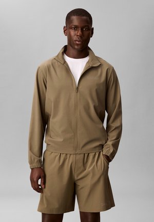 Mand iført beige lynlåstrøje og matchende shorts, stående mod en ens grå baggrund, hænderne afslappet langs siderne.