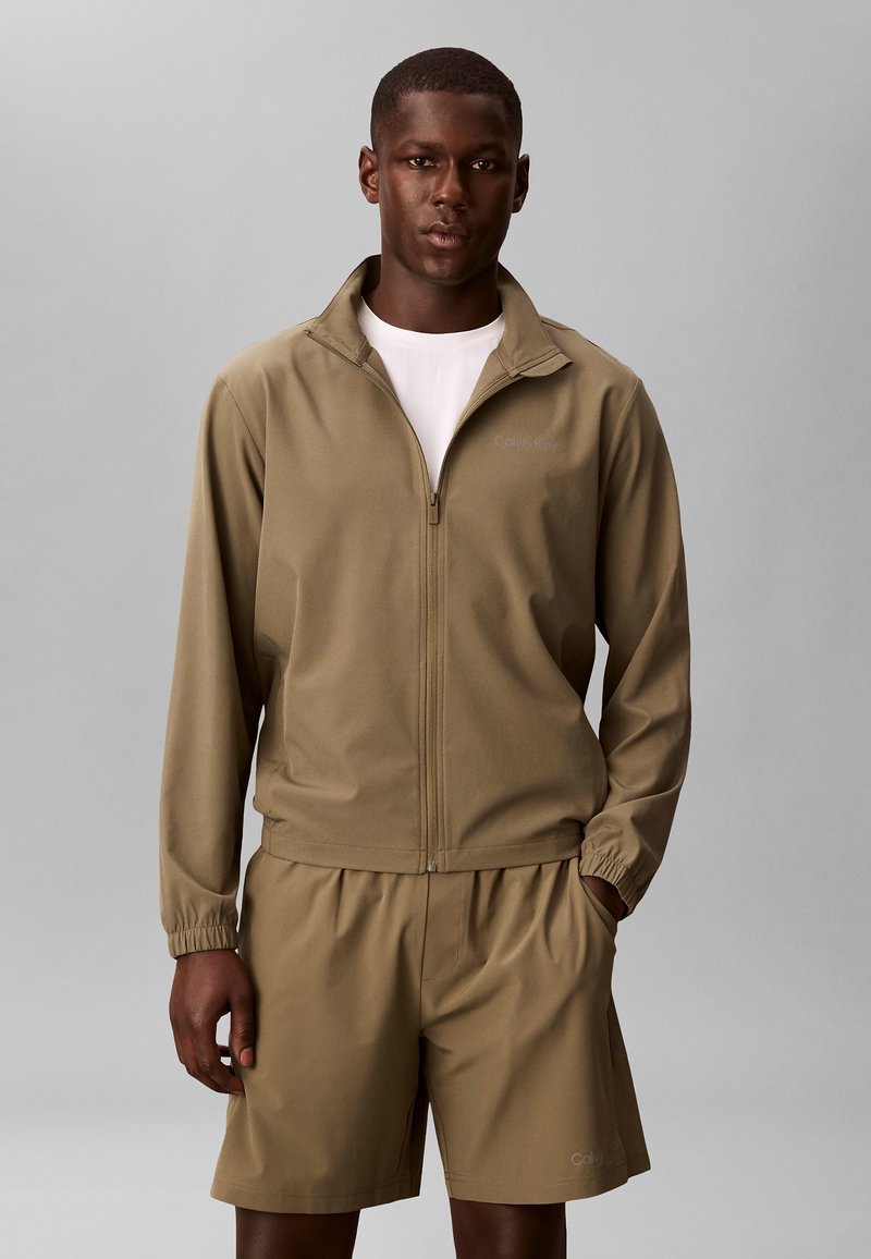 Άνδρας με μπεζ μπουφάν με φερμουάρ και αντίστοιχα σορτς, στέκεται μπροστά σε απλό γκρι φόντο, με τα χέρια χαλαρά στα πλάγια.