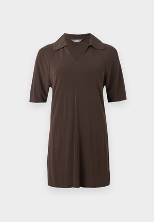 Vrouw staand, draagt een donkerbruine tuniek met korte mouwen en kraag, met bijpassende wijde broek, gecombineerd met zwarte puntige schoenen en een kettingketting.