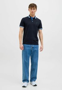 Marineblauwe poloshirt met lichtblauwe strepen op de kraag en mouwen, gecombineerd met lichtblauwe, wijde spijkerbroek. Witte sportschoenen.