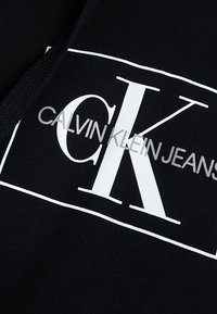 Tela negra con una textura suave que presenta un gran gráfico blanco: "CK" destacado, con "CALVIN KLEIN JEANS" debajo en texto más pequeño.