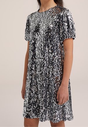 Kobieta w krótkiej sukience z krótkim rękawem do kolan, zdobionej czarno-srebrnymi cekinami, z okrągłym dekoltem, stojąca na jednolitym beżowym tle.