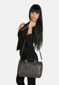 Borsa a mano in pelle marrone con texture liscia, forma arrotondata, doppio manico, tracolla rimovibile e dettaglio decorativo a chiusura.