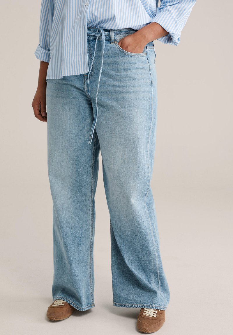 Hellblaue Jeans mit weitem Bein und Kordelzug an der Taille, mit einem verwaschenen Look und subtiler strukturierter Oberfläche, getragen mit braunen Sneakern.