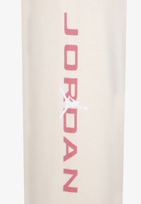 Cremfarbener Stoff mit auffälligem pinken "JORDAN"-Text und einem weißen Jumpman-Logo in der Mitte. Glatte, weiche Textur.