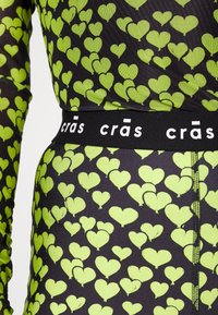Czarny top z tkaniny, zdobiony powtarzającym się wzorem w żywe zielone serca, z kontrastującą czarną elastyczną talią oznaczoną napisem "crás".