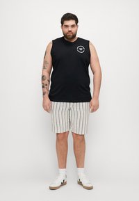 T-shirt sans manches noir avec un logo graphique, shorts rayés beige et bleu, baskets blanches avec des accents noirs et des tatouages visibles sur les bras.
