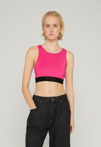 Ružová crop top vyrobená z elastického materiálu s čiernym elastickým pásom s logom, v kombinácii s čiernymi džínsy s vysokým pásom. Hladká textúra.