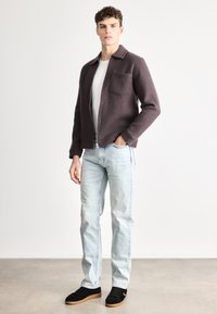 Tamnoljubičasta jakna s ovratnikom i džepom na prsima, uparena sa svijetloplavim trapericama i crnim tenisicama s gumenim đonom na neutralnoj pozadini.