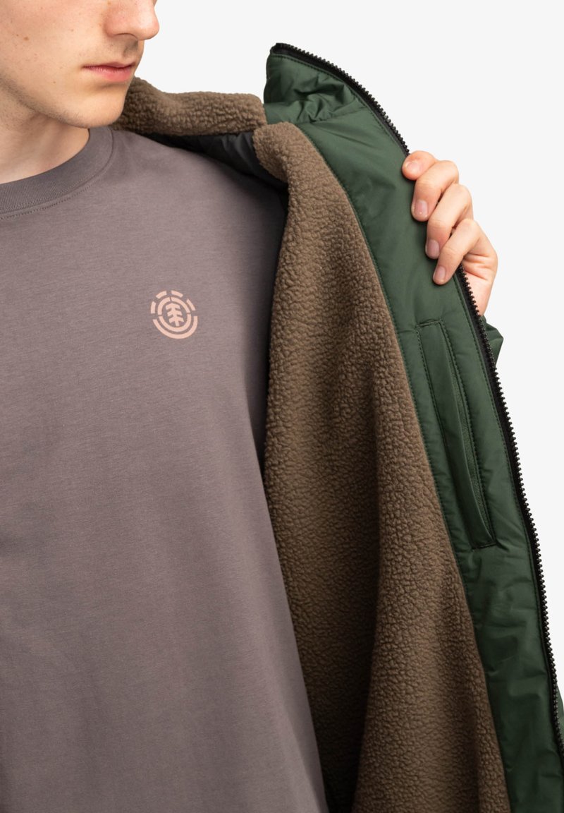 Giacca isolante verde con fodera in pile marrone, indossata aperta su una maglietta grigia con un logo grafico rosa chiaro.