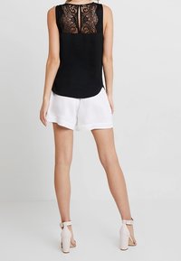 Haut sans manches noir avec détail en dentelle au dos et encolure ronde, associé à un short blanc et des talons à bride de cheville beige. Texture de tissu lisse.