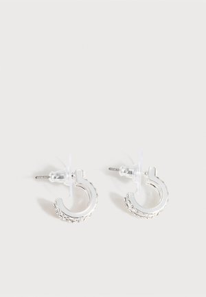 Petites boucles d'oreilles créoles ton argent avec un motif texturé le long du bord extérieur et des attaches en plastique transparent, présentées sur un fond clair.