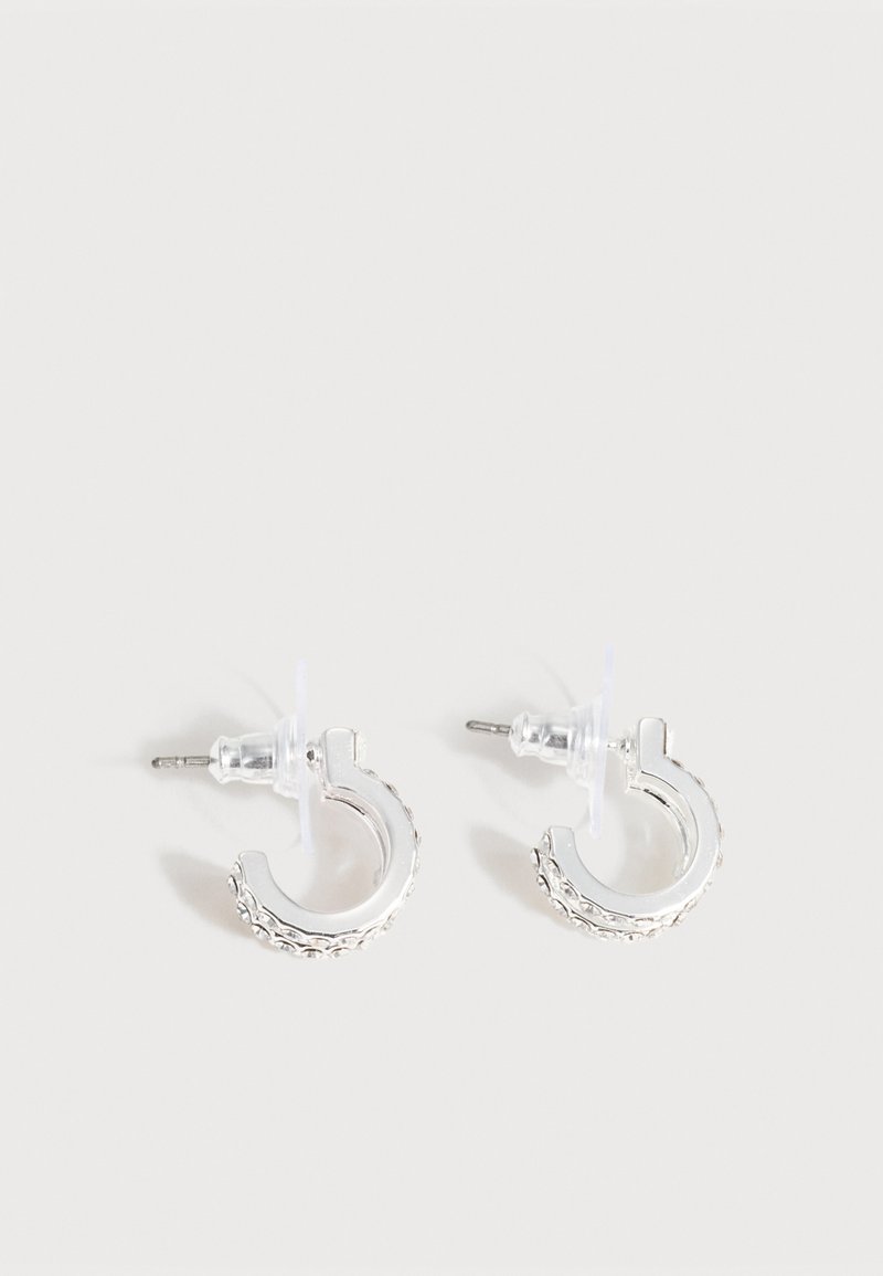 Petites boucles d'oreilles créoles ton argent avec un motif texturé le long du bord extérieur et des attaches en plastique transparent, présentées sur un fond clair.