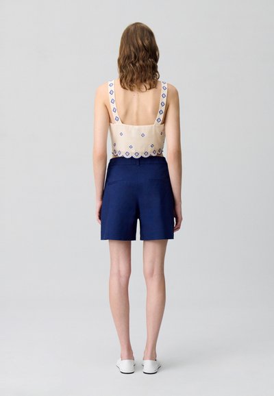 Un haut beige avec une broderie florale bleue et de larges bretelles, associé à un short marine. Des chaussures blanches à enfiler complètent le look.