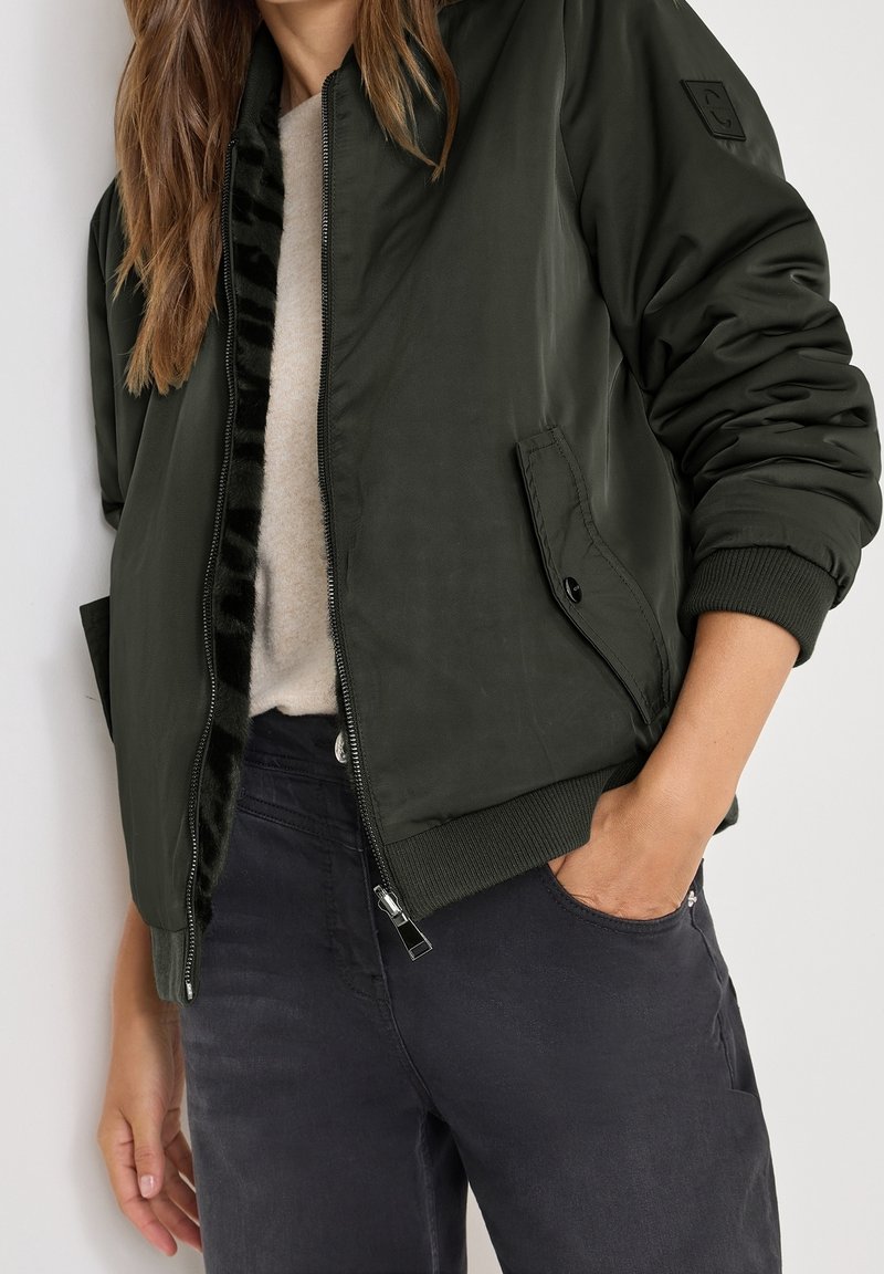 Veste bomber vert foncé avec une fermeture éclair, des poignets côtelés et une doublure noire moelleuse. Comprend une poche latérale et une texture de tissu lisse.