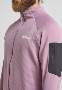 Giacca leggera e strutturata di colore viola, con colletto alto, zip frontale e una tasca con zip nera sulla manica sinistra. Logo del brand sul petto.