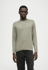 Maglione verde chiaro con scollatura rotonda e polsini a coste, abbinato a jeans grigio scuro. Tessuto morbido, vestibilità slim e dettagli di design minimi.