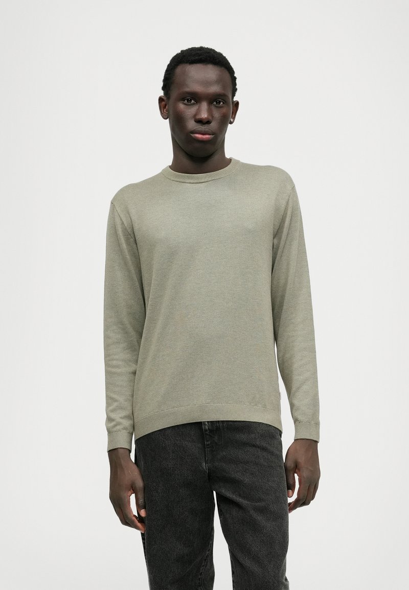 Maglione verde chiaro con scollatura rotonda e polsini a coste, abbinato a jeans grigio scuro. Tessuto morbido, vestibilità slim e dettagli di design minimi.