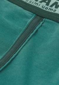 Primer plano de una tela verde azulado que muestra costuras detalladas y una cinturilla elástica gris con texto blanco parcial.