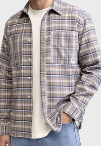 Kostkovaná košile v béžových a modrých tónech, vyrobená z flanelu. Má zapínání na knoflíky, dvě náprsní kapsy a tkanou nálepku na levé kapse.