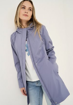 Junge Frau trägt einen helllila Kapuzenregenmantel über einem weißen Hemd mit blauem Muster und hellblauen Jeans, steht vor einem schlichten Hintergrund.